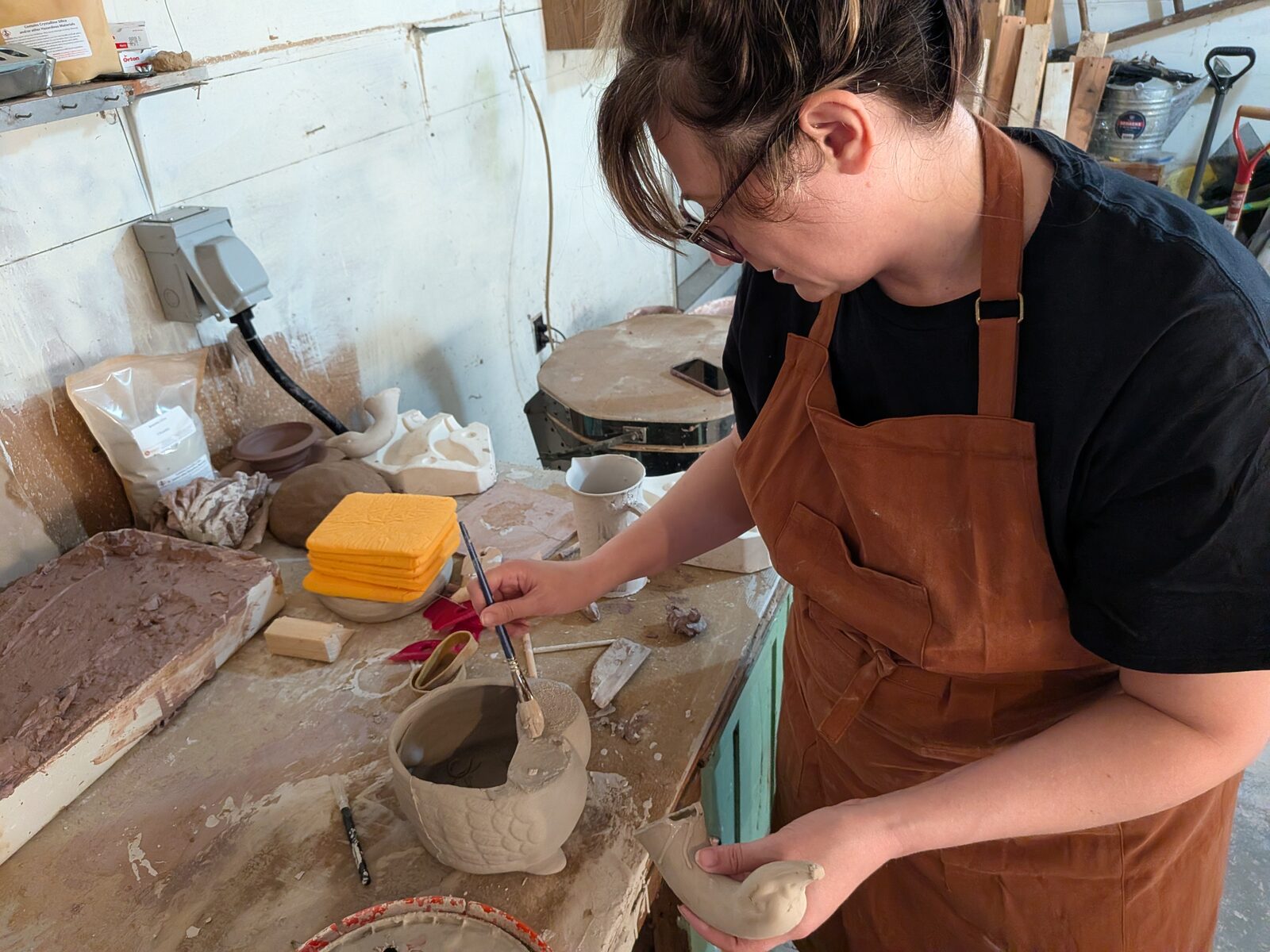 Alesha at her workbench in glasses and a brown leather apron, brushing slip into a carved clay pot while holding a small clay pig in her other hand.