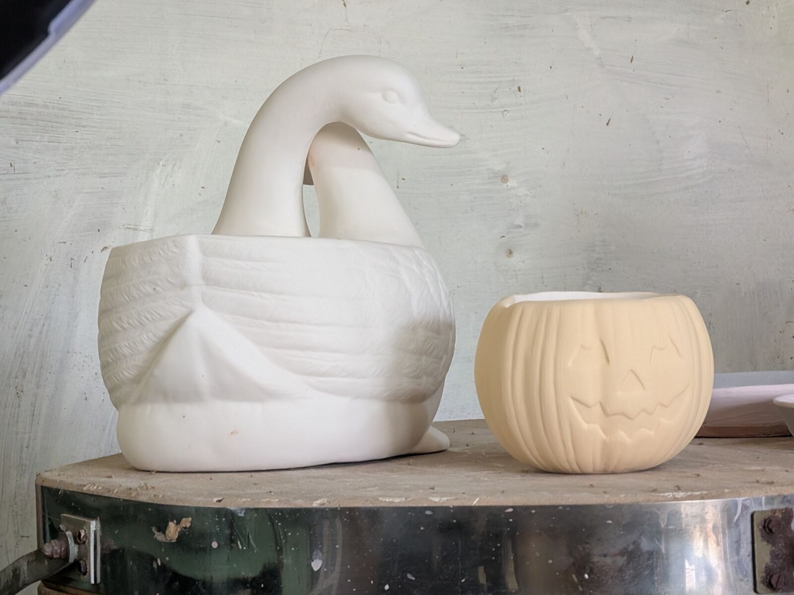 An unfired white bisque swan with a carved feather pattern, beside an unfired tan bisque jack-o'-lantern with a carved face, on a turntable.