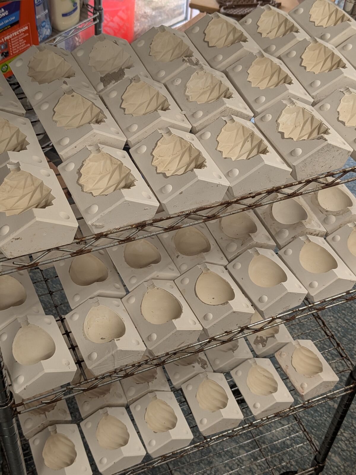 Around forty plaster slipcast moulds on a rack, some still holding carved leaf and pinecone relief, some emptied after casting.