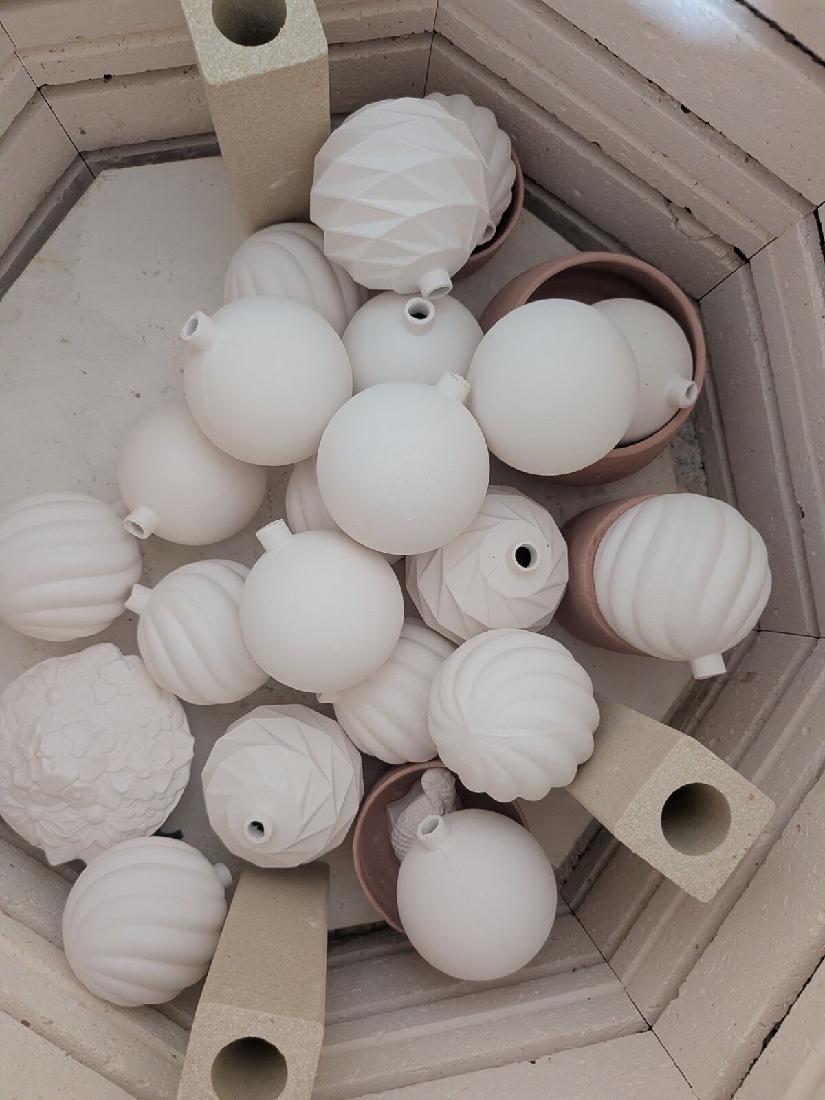 Looking down into a kiln at pale unfired ceramic ornaments with faceted, ribbed and pinecone-textured surfaces, several sitting in pink silicone moulds, separated by refractory kiln posts.