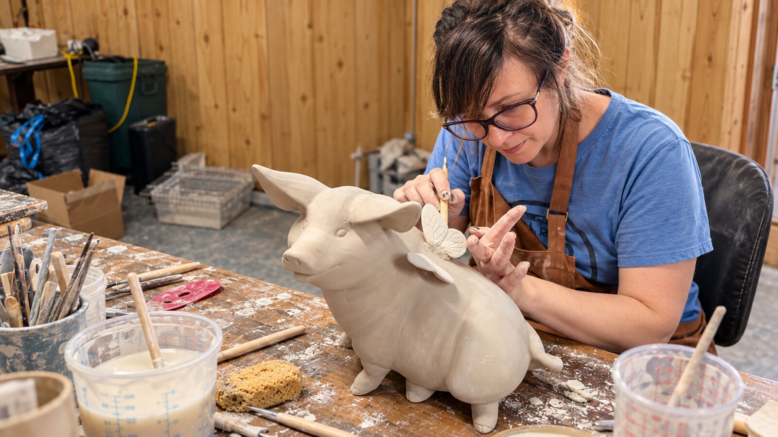Illustration of Alesha standing at a workbench holding a clay pig she has been shaping, with tools and slip nearby.