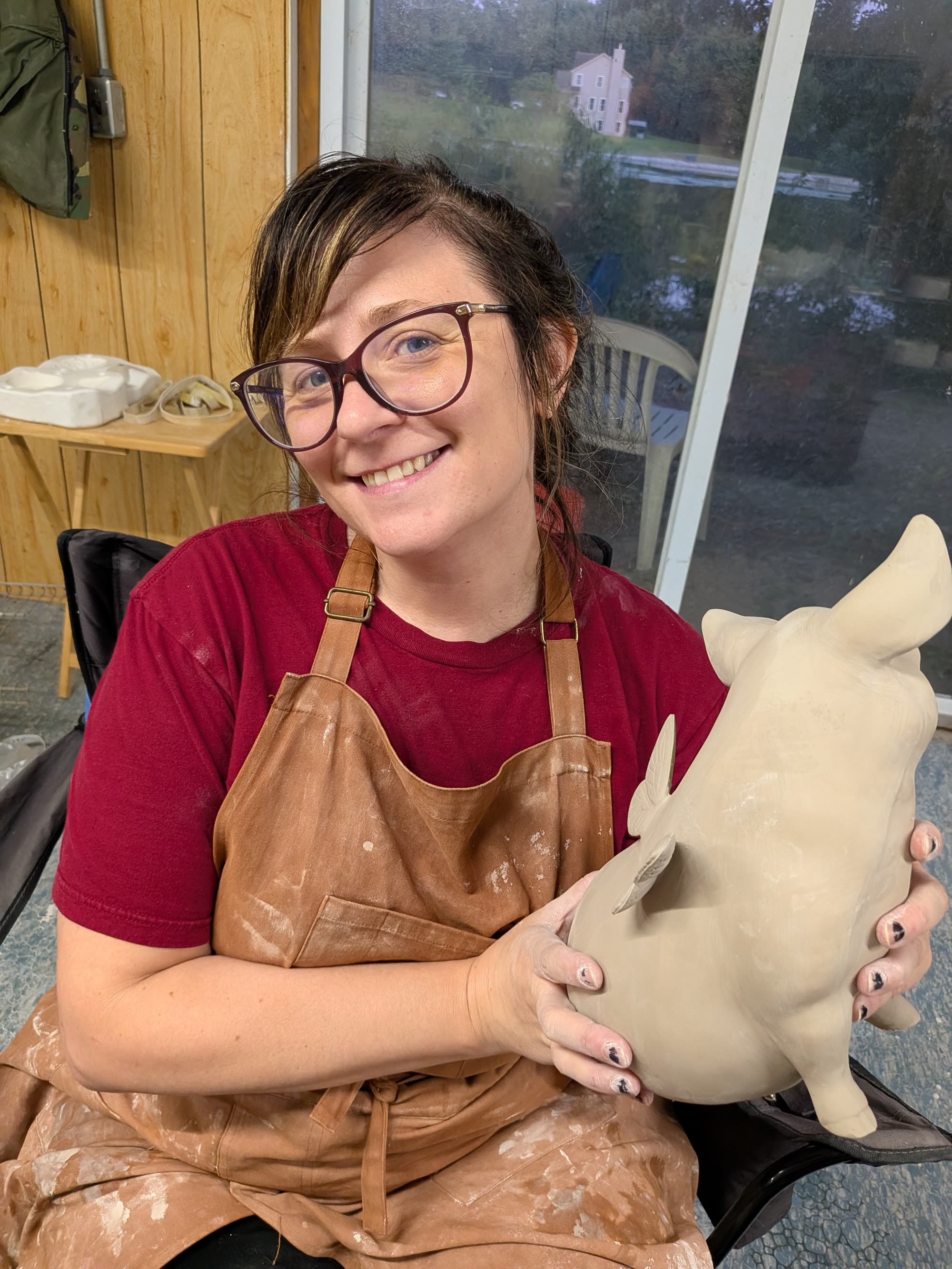 Alesha, in glasses and a clay-marked leather apron, smiles while holding a large pale clay pig with applied wings.
