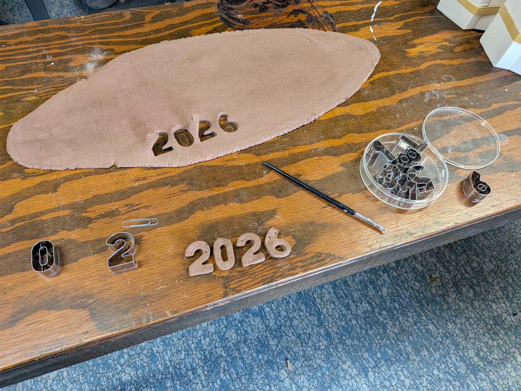 A flat oval sheet of clay on a wooden table, with the numerals 0 2 2 6 cut out and laid beside it and a craft knife alongside.