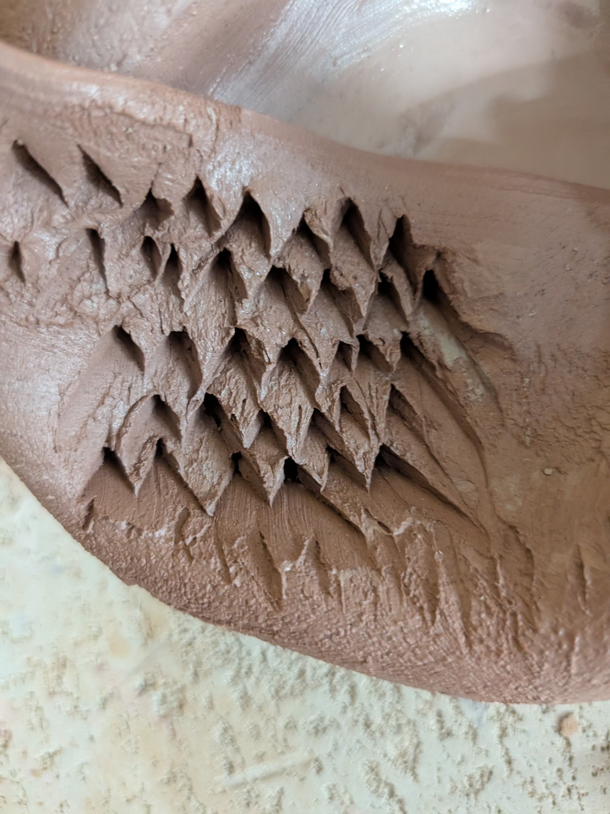 A close view of pale unfired clay carved with a repeating pattern of overlapping carved scales.